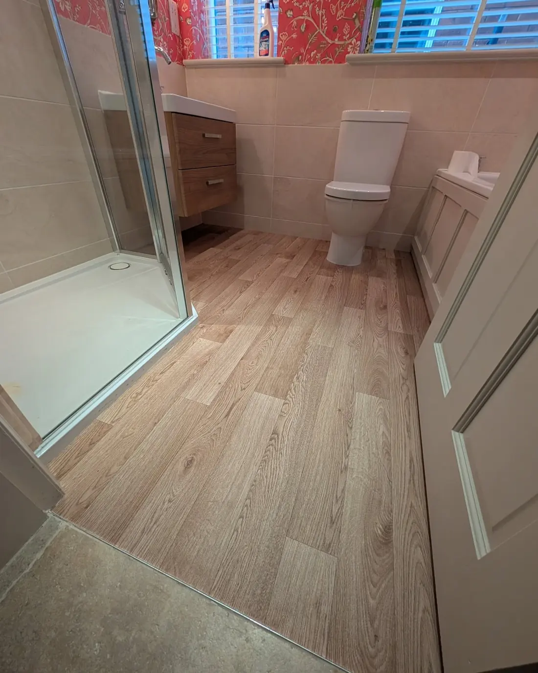 The same bathroom finished with pale wood effect plank flooring, red patterned wallpaper unchanged.