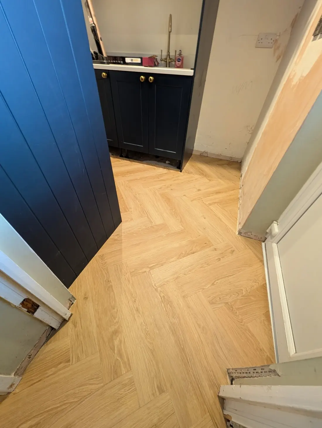 The same kitchen finished with natural oak herringbone LVT running past the navy shaker units and their brass handles.