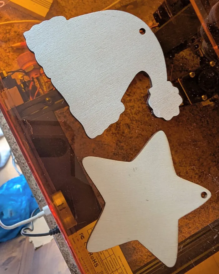 Two blank wooden shapes, a Santa hat and a star, each with a small hanging hole, waiting on her laser's orange lid.
