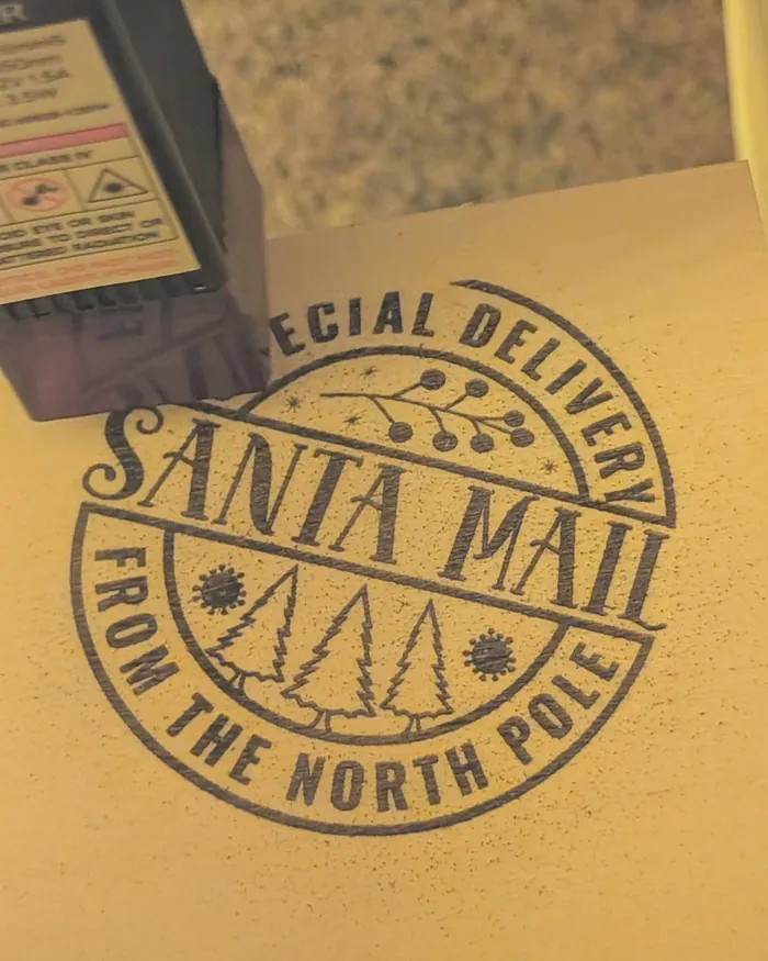 Her laser engraver's head beside a round 'Santa Mail, Special Delivery from the North Pole' stamp it has just engraved into the wood.