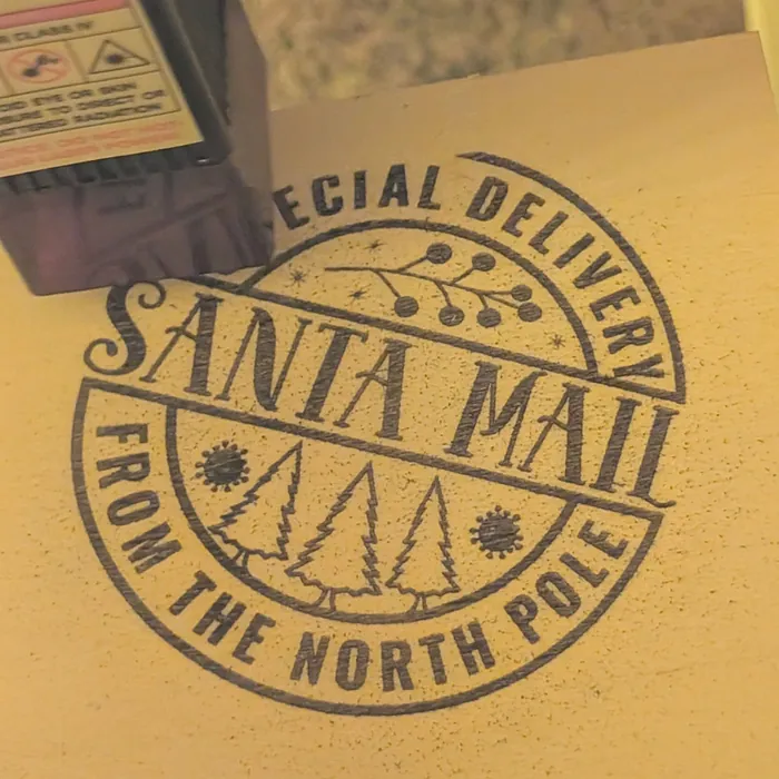 A round 'Santa Mail, Special Delivery from the North Pole' stamp engraved into pale wood under her laser's head.