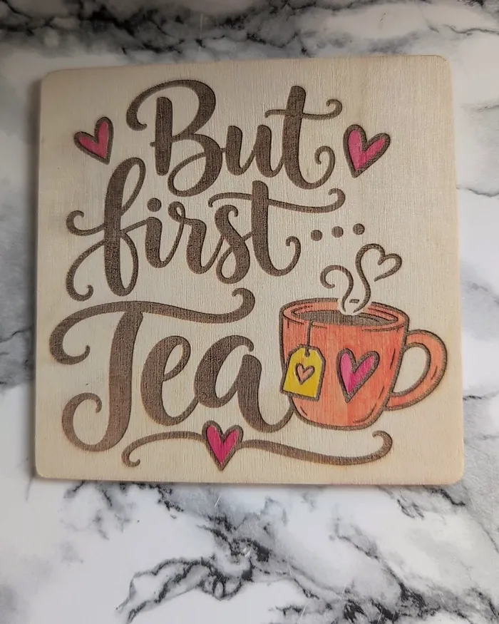 A square wooden coaster engraved 'But first... Tea' with hand-painted pink hearts, an orange teacup and a yellow tea tag.