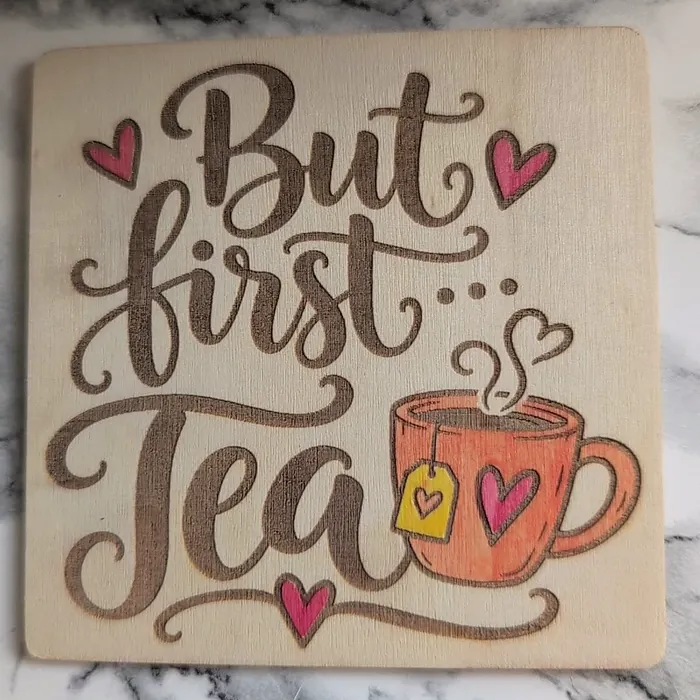 A square coaster engraved 'But first... Tea' with hand-painted pink hearts and an orange teacup.