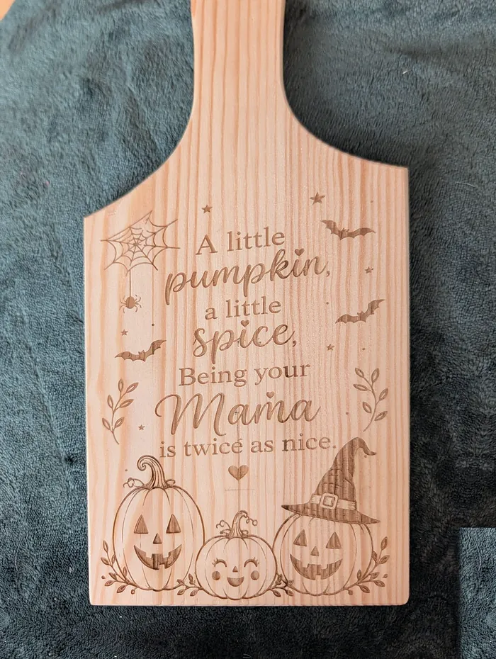 Her pine chopping board engraved 'A little pumpkin, a little spice, Being your Mama is twice as nice' with jack-o'-lanterns, on a dark blanket.
