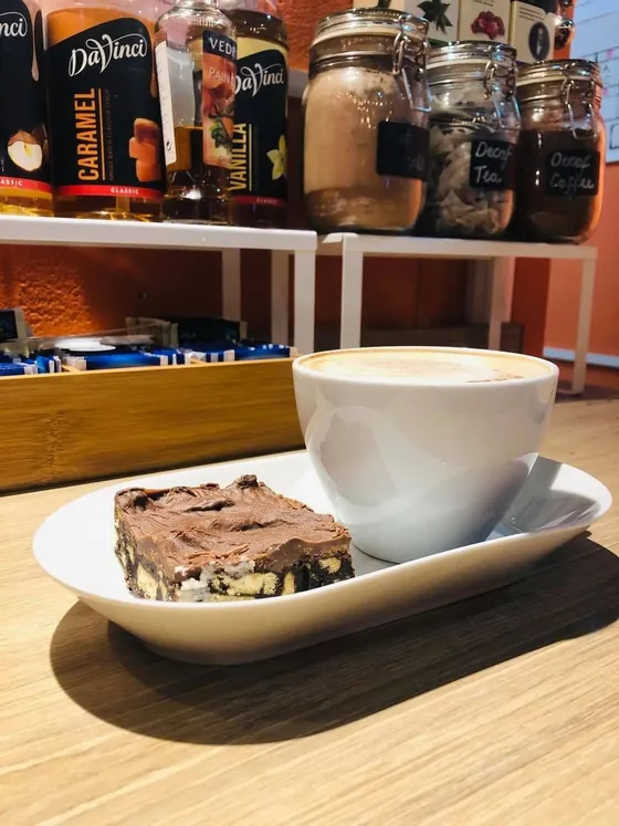 A slice of chocolate cake beside a cappuccino, with syrup bottles and storage jars on the shelf behind.