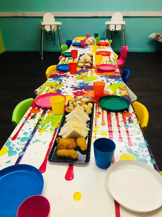 A long party table with plates, coloured cups and platters of food, rainbow chairs down both sides.