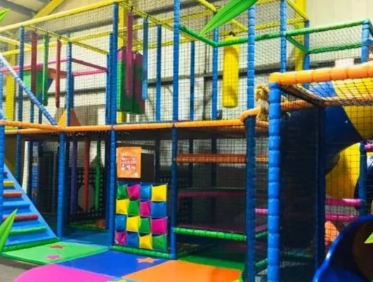 The three level play frame from the front, with orange and blue rails, a yellow tube slide and rainbow padded floor mats.
