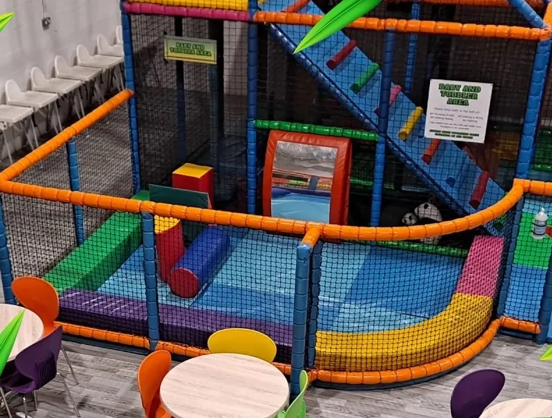The soft play frame seen from the cafe side, with orange tube rails, blue netting and a green slide across several levels.