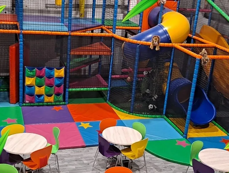 The full play frame with ball pit, yellow and blue tube slides and orange monkey bar rails, with cafe tables alongside.