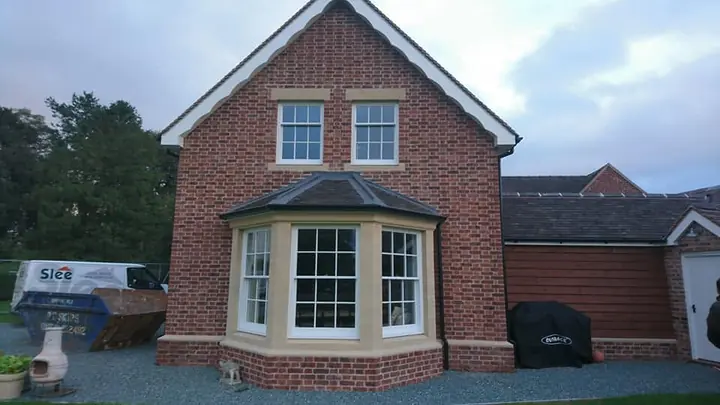 A finished red-brick house with a white-trimmed gable, sash windows and a bay window under a small hipped roof, beside a lower brick outbuilding with a dark tiled roof.