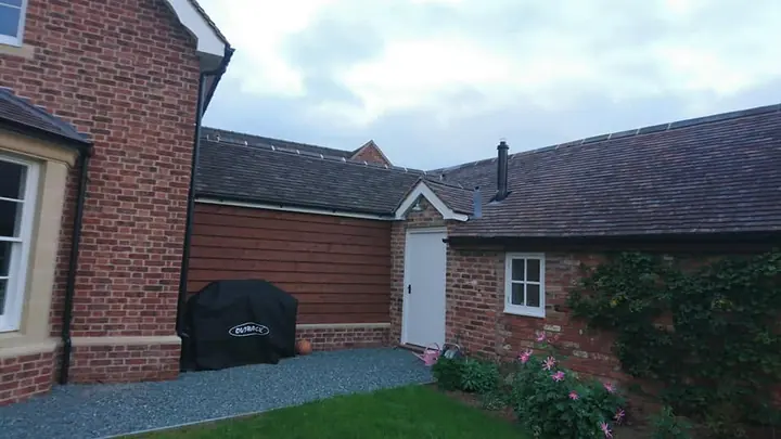 The side of a red-brick house joined to a range of single-storey outbuildings with dark tiled roofs, black gutters and white doors, beside a lawn and climbing plants.
