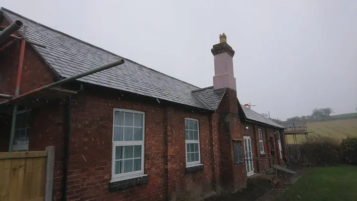 A long red-brick building with a slate roof, a tall chimney and white sash-style windows, with scaffolding at one end under a grey sky.