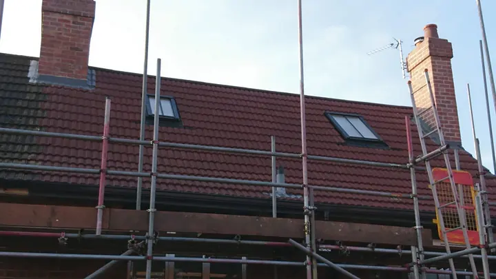 A red tiled roof with two roof windows and two brick chimneys, surrounded by scaffolding and a ladder under a clear sky.