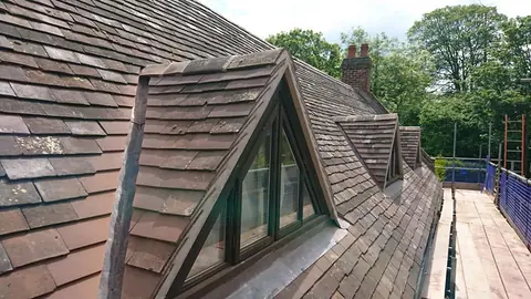 Close-up of a small triangular dormer with a timber-framed window in an old plain-tile roof, with a scaffold walkway and blue safety netting alongside.