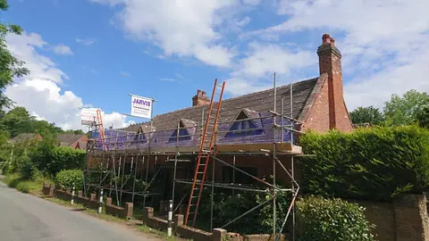 A brick cottage with a tall chimney and small triangular dormers in its tiled roof, wrapped in scaffolding with blue safety netting and a ladder, beside a country lane.
