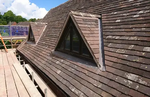 Two small triangular dormers in a weathered plain-tile roof, with a scaffold walkway, blue safety netting and an orange ladder alongside.
