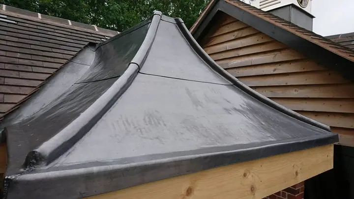Close-up of the curved canopy roof: a dark sheet covering with a rolled ridge joint and a rolled edge over a timber beam, beside a weatherboarded gable.