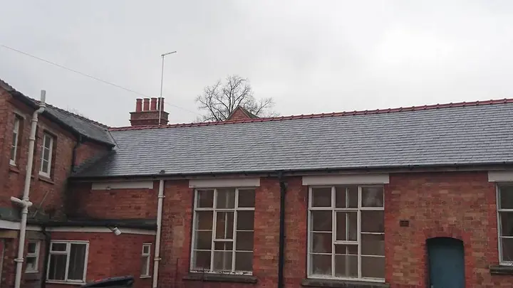A long red-brick building with a slate roof edged with red ridge tiles, white-framed windows and a blue door, under a grey sky.