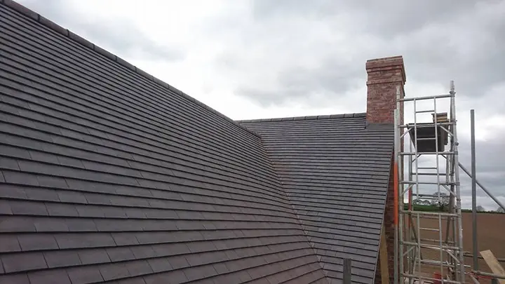 Dark plain tiles on two new roof slopes meeting at an angle, with a brick chimney and a scaffold tower beside it.