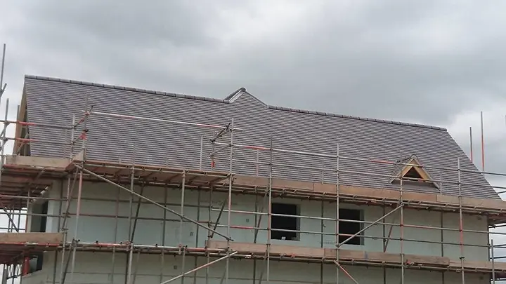 A large new house under scaffolding with a roof of dark plain tiles and a small gabled vent, its walls still unfinished, under a grey sky.