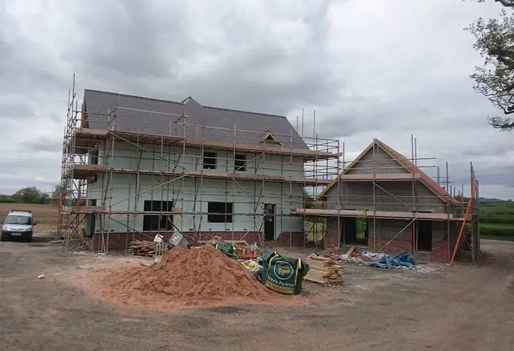 A large house under scaffolding in open farmland, with a new tiled roof and a smaller gabled wing, and a van and a heap of sand on the site in front.