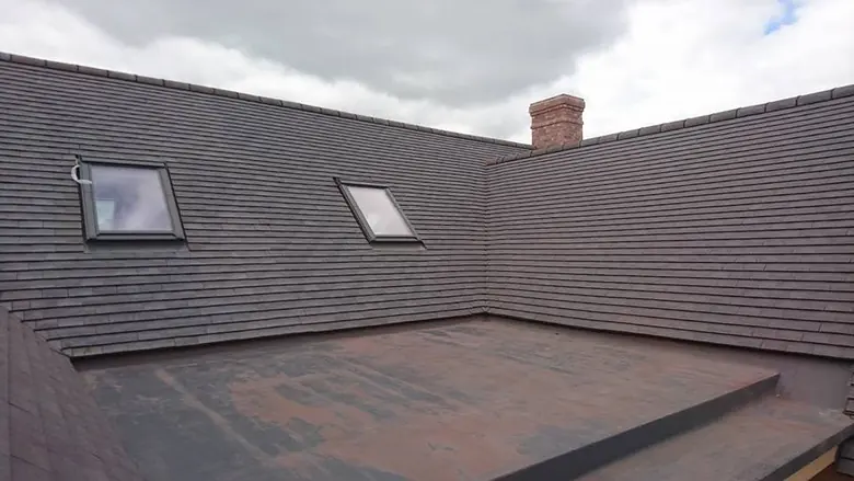 The same flat roof from another corner, framed by tiled slopes with two roof windows and a brick chimney.