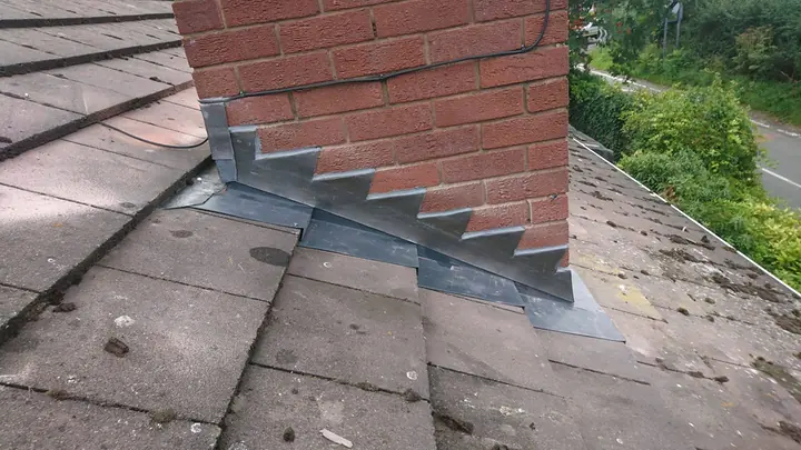 New stepped lead flashing dressed down the side of a red-brick chimney where it meets a tiled roof, with a road and hedges below.