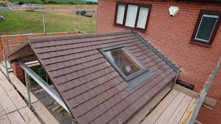 A plain-tiled lean-to roof with a roof window, against a red-brick wall, with scaffold boards on both sides and fields beyond.