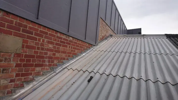 A grey corrugated sheet roof meeting a red-brick wall, with new lead step flashing running up the wall along the join.