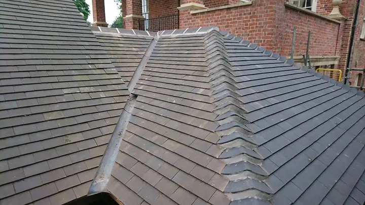 Plain tiles on a roof where slopes meet, with a grey lead roll down one hip and shaped hip tiles down the other, beside a red-brick building.