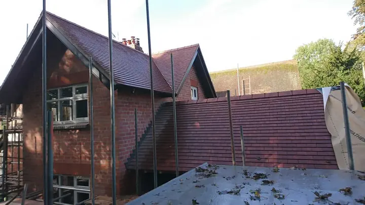 New red-brown plain tiles on a lower roof beside a tall red-brick gable, seen from a scaffold deck scattered with leaves.