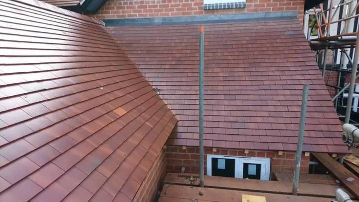 Red-brown plain tiles on two new roof slopes against a red-brick wall, with a small window below and scaffold boards around the eaves.