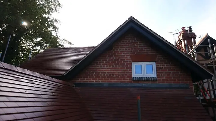 A red-brick gable with a dark bargeboard and a small white window, above a newly tiled red-brown plain-tile roof, with scaffolding and trees beyond.