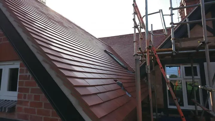 A steep slope of new red-brown plain tiles running down beside a dark bargeboard, with scaffolding and a ladder against the house.