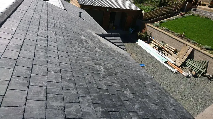 Looking down a new slate roof, course after course, towards a gravel yard with timber stacked beside a sloping lawn.