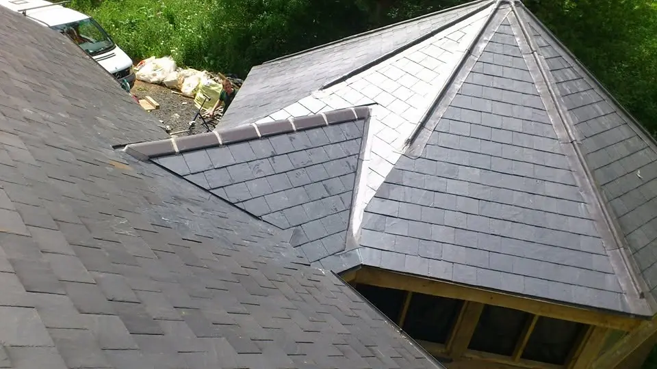 Looking down on a freshly slated hipped roof with lead-covered hips, where it meets an older slate roof above a timber-framed opening, with a van and builders' bags in the yard beyond.