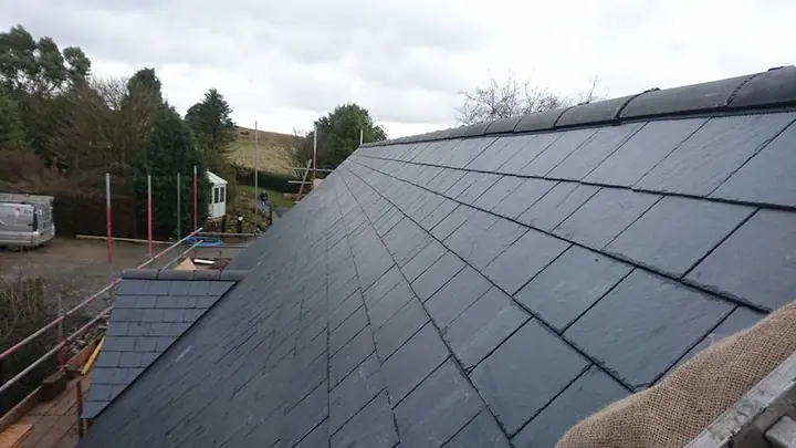 Close-up of a new slate roof with dark ridge tiles, seen from the scaffold, with a yard, a van and hills beyond.