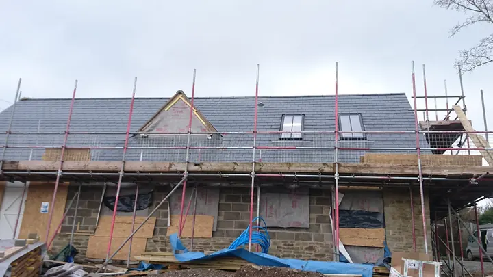 A stone house under scaffolding with a freshly laid slate roof, a gabled dormer still in membrane and two roof windows, its window openings boarded and a blue tarpaulin in front.