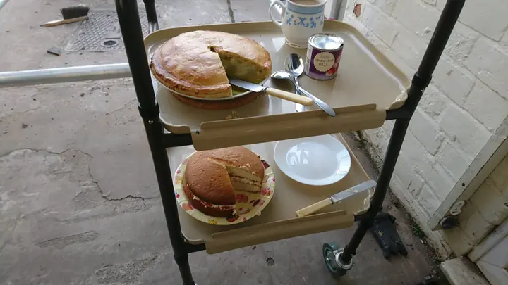 A two-tier trolley set with two sponge cakes, a jug, a tin, plates and knives, left out beside a painted brick wall.