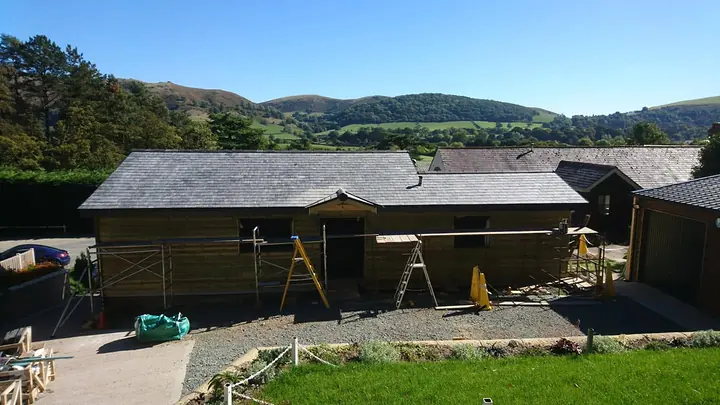 Timber-clad single-storey buildings with new slate roofs and ladders against the eaves, in a gravel yard with green hills behind under a blue sky.