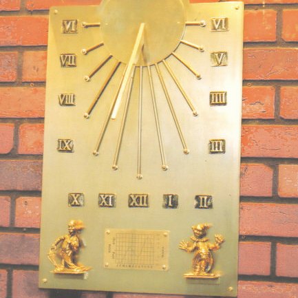 The Auguste south-facing wall dial mounted on a red brick wall: a tall brass plate with a raised sun disc and projecting gnomon at the top, rays radiating out to ball tips, Roman numeral hours set around and beneath them, and at the foot an engraved Equation of Time chart flanked by two small cast figures, one carrying an umbrella and one with its arms raised.