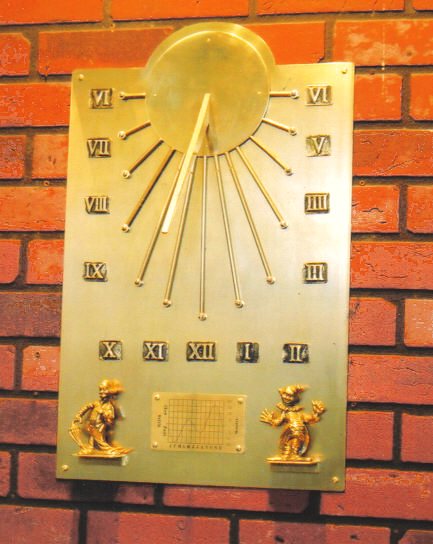 The Auguste south-facing wall dial mounted on a red brick wall: a tall brass plate with a raised sun disc and projecting gnomon at the top, rays radiating out to ball tips, Roman numeral hours set around and beneath them, and at the foot an engraved Equation of Time chart flanked by two small cast figures, one carrying an umbrella and one with its arms raised.