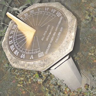 A Jacobean dial photographed from above in a garden, its brass plate carrying Roman numerals around the rim and a personal inscription engraved across the face beside the hour lines, set into an octagonal stone base surrounded by ivy and low planting.