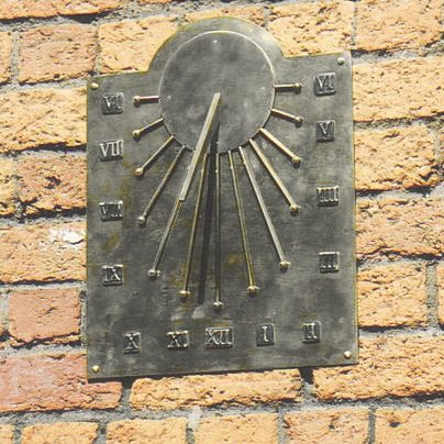 The Cameo south-facing wall dial mounted on a buff brick wall: an arched brass plate toned green with age, with a raised sun disc and projecting gnomon at the top, rays radiating out to ball tips, and Roman numeral hours running from VI on the left, through XII at the foot, to VI on the right.