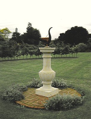 A Cygnus swan sundial on a pale Linford pedestal standing on a brick paved circle in a large lawn, with an avenue of trees behind it.