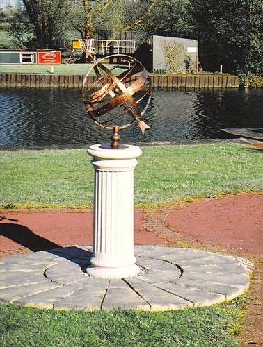 An Orbis armillary on a fluted Doric column pedestal, standing on a circle of block paving cut into a lawn beside water.