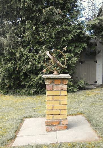 A Draco armillary mounted on a yellow brick pier set on a concrete pad, with conifers and a house behind it.