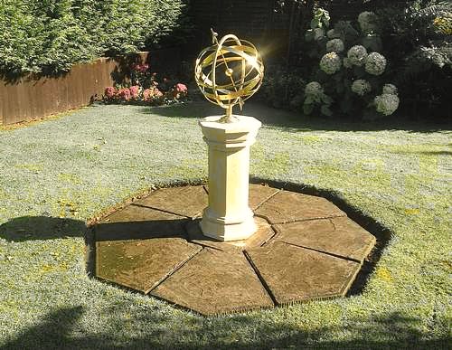 An Ecliptic armillary on a pale stone Upton pedestal, standing at the centre of an octagon of paving cut into a lawn, with hydrangeas and a fence behind it.