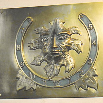 The Green Man wall dial: a landscape brass plate carrying a foliate face cast in high relief, its hair and beard formed from leaves, ringed by a horseshoe-shaped band finished in green verdigris and numbered in Arabic hours from 7 on the left, round through 12 at the foot, to 5 on the right, with leaf sprays at the lower corners.