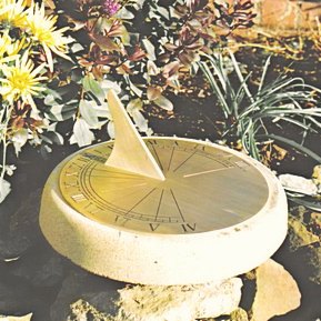 The round Jacobean brass sundial seated on a pale stone plinth in a rockery, its slim brass gnomon throwing a shadow across the engraved hour lines and numerals, with yellow chrysanthemums and grasses growing behind.